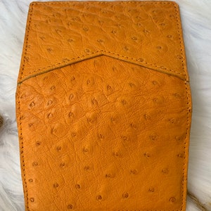 Puede incluir: Una cartera de cuero marr&oacute;n con un patr&oacute;n texturizado de piel de avestruz. La cartera tiene un cierre de solapa y un solo detalle de costura.