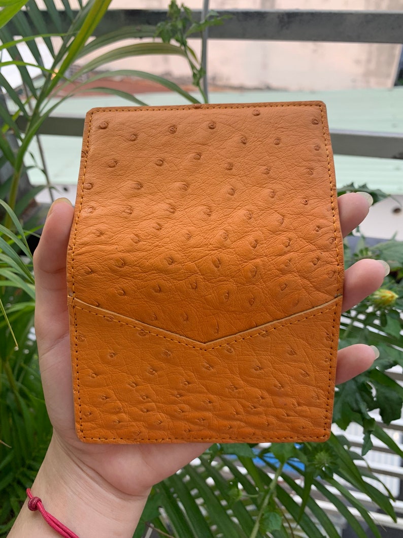 Puede incluir: Una peque&ntilde;a cartera de cuero naranja con un patr&oacute;n texturizado de piel de avestruz. La cartera tiene un cierre de solapa y un solo bolsillo en la parte delantera.