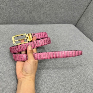 May include: Pink belt with a textured, reptile-like pattern. The belt has a gold-colored buckle. The belt is shown in a folded position.
