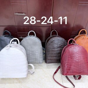 Può includere: Una collezione di sei zaini con texture a stampa coccodrillo, presentati in nero, bianco, grigio, marrone scuro, bordeaux e arancione. Ogni zaino presenta una parte superiore arrotondata, un manico e due spallacci. Il testo 28-24-11 è visibile.