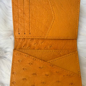 Puede incluir: Una cartera de cuero amarilla con m&uacute;ltiples ranuras para tarjetas y un bolsillo grande. La cartera est&aacute; hecha de cuero de avestruz y tiene una superficie texturizada.