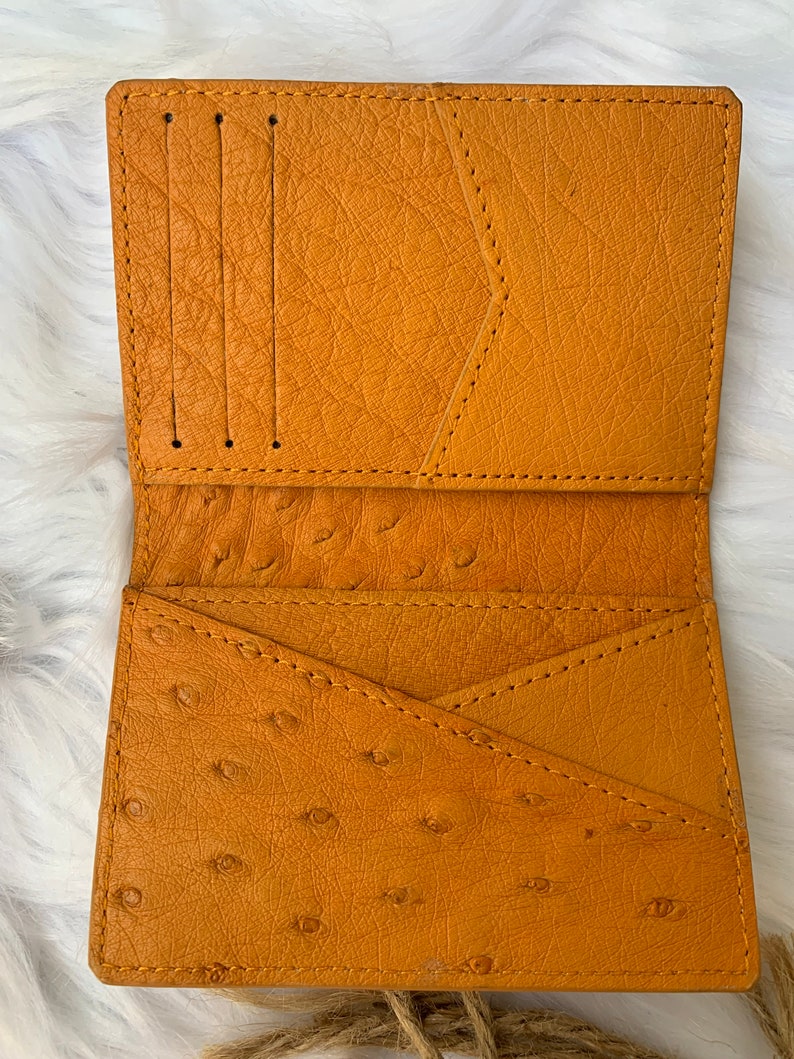 Puede incluir: Una cartera de cuero amarilla con m&uacute;ltiples ranuras para tarjetas y un bolsillo grande. La cartera est&aacute; hecha de cuero de avestruz y tiene una superficie texturizada.