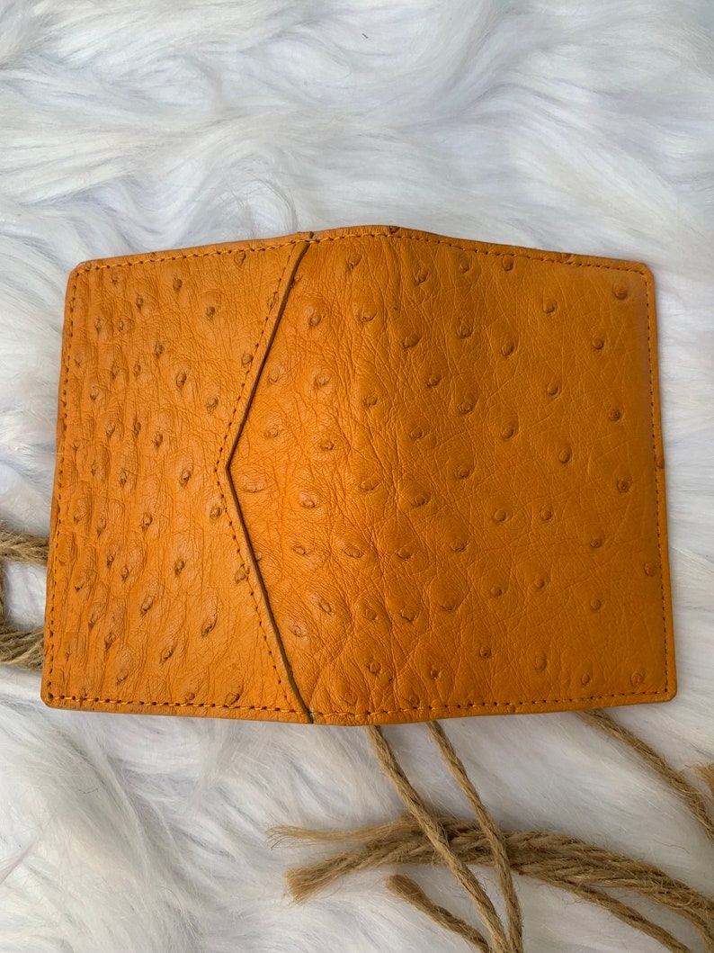 Puede incluir: Una peque&ntilde;a cartera de cuero amarilla mostaza con un patr&oacute;n de avestruz texturizado. La cartera tiene un cierre de solapa y est&aacute; hecha de cuero de alta calidad.