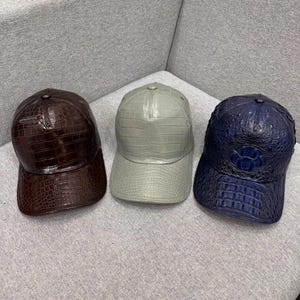 May include: Three baseball caps in a row, each with a textured, crocodile-like pattern. The caps are in shades of brown, gray, and navy blue. The caps are sitting on a gray surface.