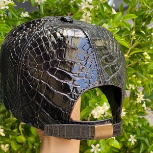 May include: Black baseball cap with a glossy, textured crocodile skin pattern. The cap features a curved brim, a small button on top, and an adjustable strap with a brass-colored buckle. The cap is a dark, shiny black.