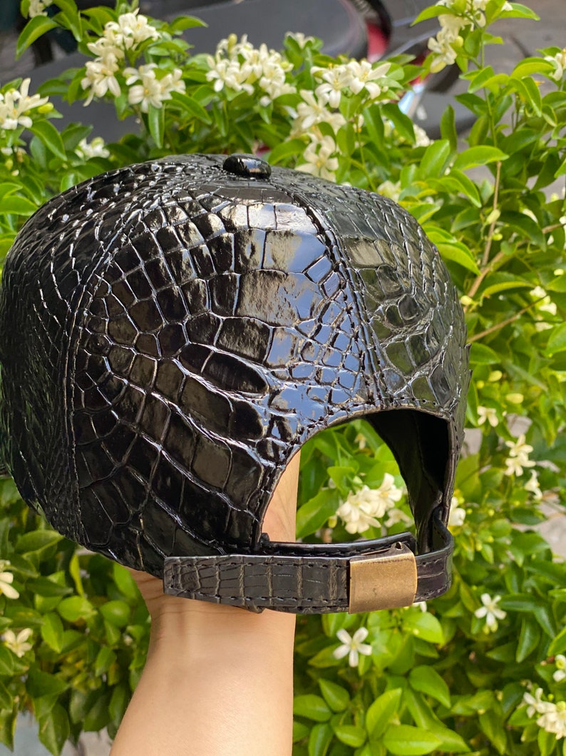 May include: Black baseball cap with a glossy, textured crocodile skin pattern. The cap features a curved brim, a small button on top, and an adjustable strap with a brass-colored buckle. The cap is a dark, shiny black.