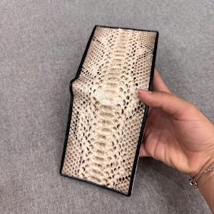 May include: A rectangular wallet with a snakeskin pattern in cream and brown shades. The wallet has a black border and a folded design. The image shows the wallet against a grey background.