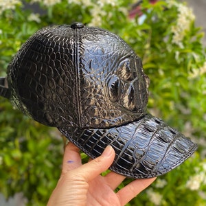 May include: A black baseball cap with a glossy, textured finish, resembling crocodile skin. The cap features a curved brim and a small button on top. The cap is held up against a backdrop of green foliage.