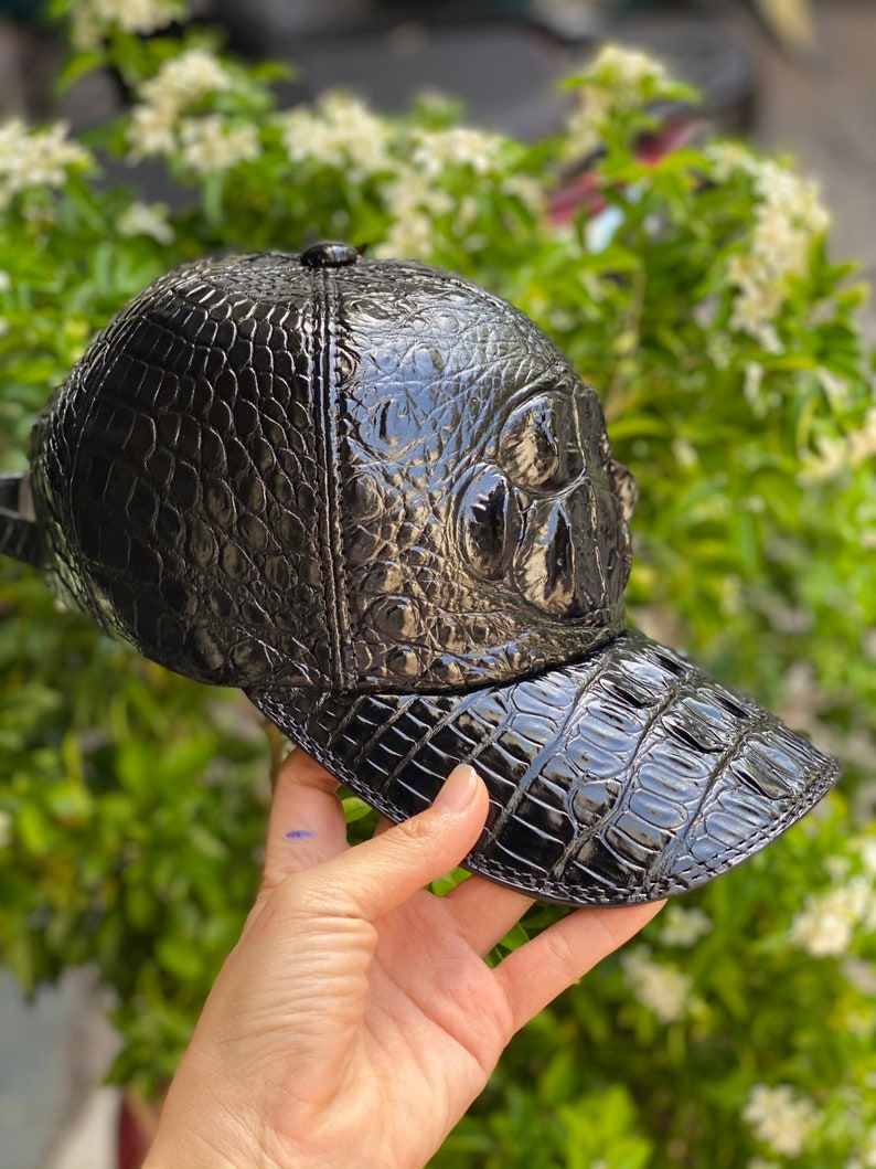 May include: A black baseball cap with a glossy, textured finish, resembling crocodile skin. The cap features a curved brim and a small button on top. The cap is held up against a backdrop of green foliage.