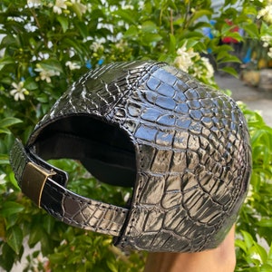 May include: Black baseball cap with a textured, crocodile-like pattern. The cap features a curved brim and an adjustable strap with a gold-colored metal buckle. The cap is made of a shiny material.
