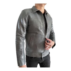 May include: A gray leather jacket with a crocodile skin texture. The jacket has a collared neckline, a zipper closure, and a buttoned waist. The sleeves are long, and the jacket appears to be a medium weight.