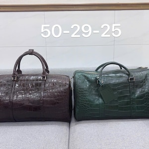 May include: Two leather travel bags with a crocodile skin texture. One bag is dark brown, the other is dark green. Both have top handles and metal hardware. The bags are on a gray sofa. The text "50-29-25" is above the bags.