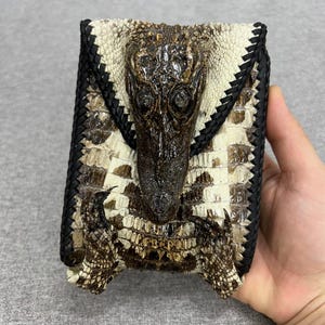Puede incluir: Una cartera artesanal con cabeza de cocodrilo y cuero texturizado. La cartera tiene un borde trenzado negro y un cierre de solapa. La paleta de colores incluye tonos de marrón, crema y negro.