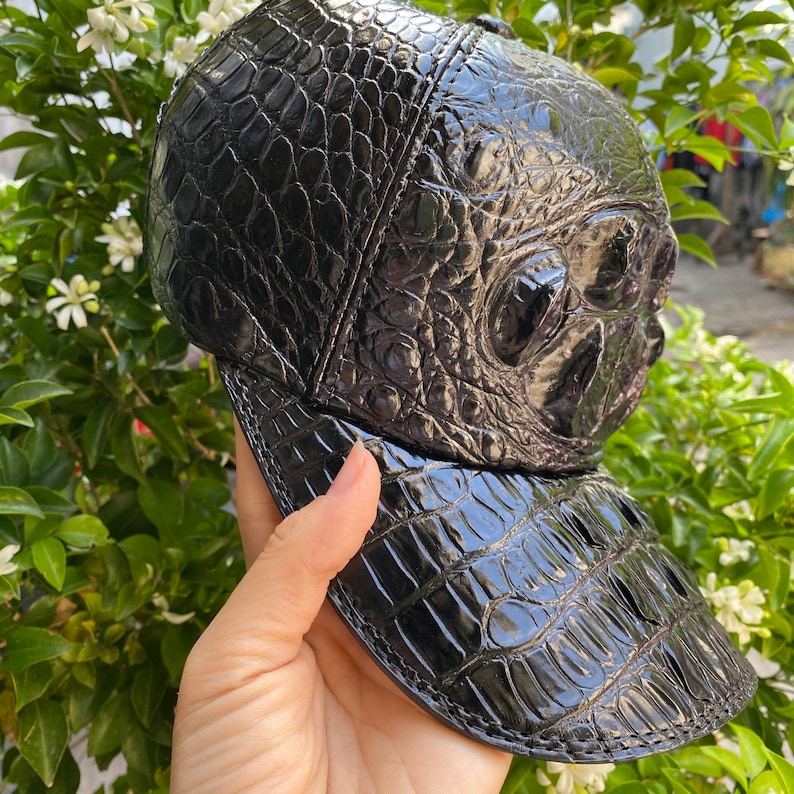 May include: A black baseball cap with a textured, crocodile-like pattern. The cap features a curved brim and a structured crown, showcasing a glossy finish. The design is a unique accessory.