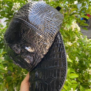May include: A black baseball cap with a textured, glossy finish, resembling crocodile skin. The cap features a curved brim and a structured crown. The background includes green foliage and white flowers.