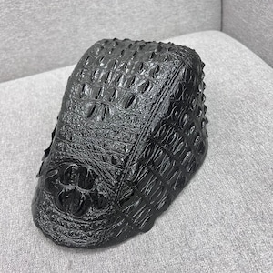 Könnte beinhalten: Eine schwarze Schiebermütze mit glänzender, strukturierter Oberfläche, die das Aussehen von Krokodilleder nachahmt. Die Mütze hat eine strukturierte, abgerundete Krone und einen kurzen, festen Schirm mit einem detaillierten, erhabenen Schuppenmuster.