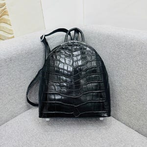 Puede incluir: Mochila negra con textura de piel de cocodrilo. La mochila tiene una parte superior curvada, un cierre de cremallera y dos correas ajustables para los hombros. Tiene un asa superior y pequeños pies blancos en la parte inferior.