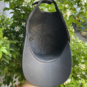 May include: Black leather baseball cap with a curved brim and adjustable strap. The cap has a stitched design and a button on top. The interior is black. The cap is shown against a background of green leaves and white flowers.