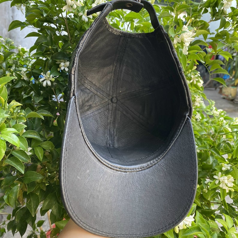 May include: Black leather baseball cap with a curved brim and adjustable strap. The cap has a stitched design and a button on top. The interior is black. The cap is shown against a background of green leaves and white flowers.