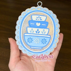 May include: An oval-shaped ornament featuring a stack of four blue and white mixing bowls with floral and heart patterns. The design is framed by a scalloped blue border and a glittery silver background. The text Catalyst fragrance co. is visible.