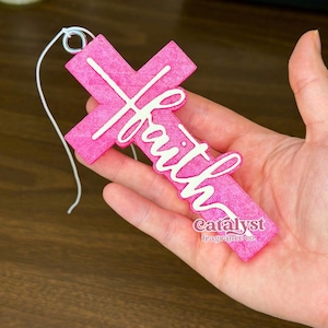 May include: A bright pink cross-shaped ornament featuring the word faith in white script lettering across the center. The textured pink surface has a small hole at the top with a white string attached for hanging. The text catalyst fragrance co is visible.
