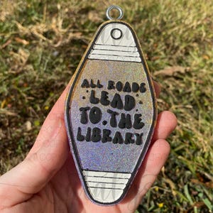 May include: A glittery, motel-style keychain with the text "ALL ROADS LEAD TO THE LIBRARY." The keychain has a gold border, white accents, and a silver metal ring for attachment. The background is blurred.