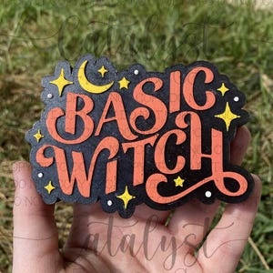 May include: A black and orange Basic Witch sign with a crescent moon and stars. The sign is held in a hand against a green background. The text is in a bold, stylized font, perfect for Halloween decor.