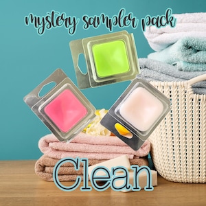 May include: A "mystery sampler pack" with three square wax melts in pink, green, and white, displayed against a backdrop of folded towels and a basket. The word "Clean" is written in a teal font at the bottom. The text "mystery sampler pack" is written in a teal font at the top.