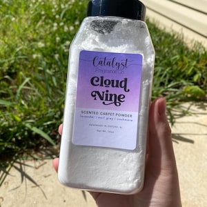 May include: A white plastic jar with a black lid filled with white powder. The jar has a label that says "Catalyst Fragrance Co. Cloud Nine Scented Carpet Powder lavender earl grey | cashmere Handmade in Oxford, AL Net Wt. 16oz"