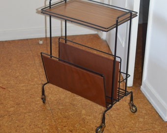 Puede incluir: Un revistero de metal negro con un estante superior de madera y portarevistas de cuero marrón. El revistero tiene cuatro ruedas y está sobre un suelo de corcho.