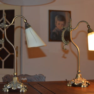 May include: Two antique brass table lamps with white cone-shaped lampshades. The lamps have ornate bases and decorative arms.