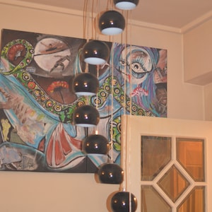 May include: A black chandelier with ten round black pendant lights hanging from a ceiling. The chandelier is in front of a colorful abstract painting.