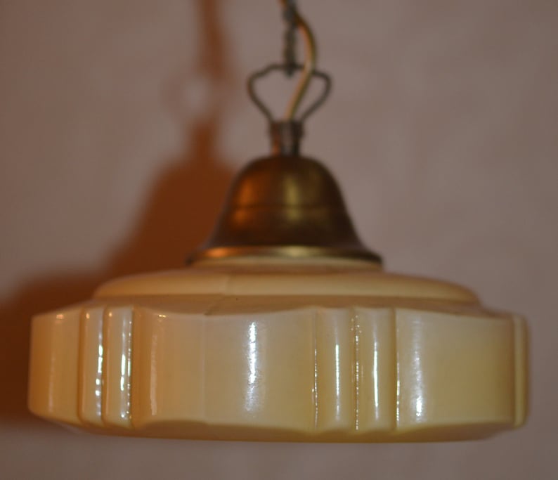 K&ouml;nnte beinhalten: Eine Vintage-Pendelleuchte mit einem cremefarbenen Glasschirm und einem messingfarbenen, glockenf&ouml;rmigen Oberteil. Der Schirm hat einen gewellten Rand und vertikale Linien.