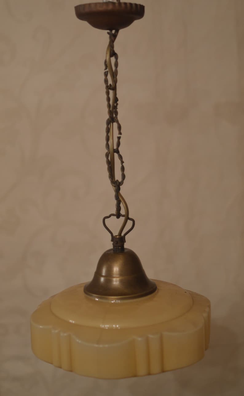 K&ouml;nnte beinhalten: Eine antike Messing-Pendelleuchte mit einem cremefarbenen Glasschirm. Der Schirm hat einen gewellten Rand und ein dekoratives Muster.