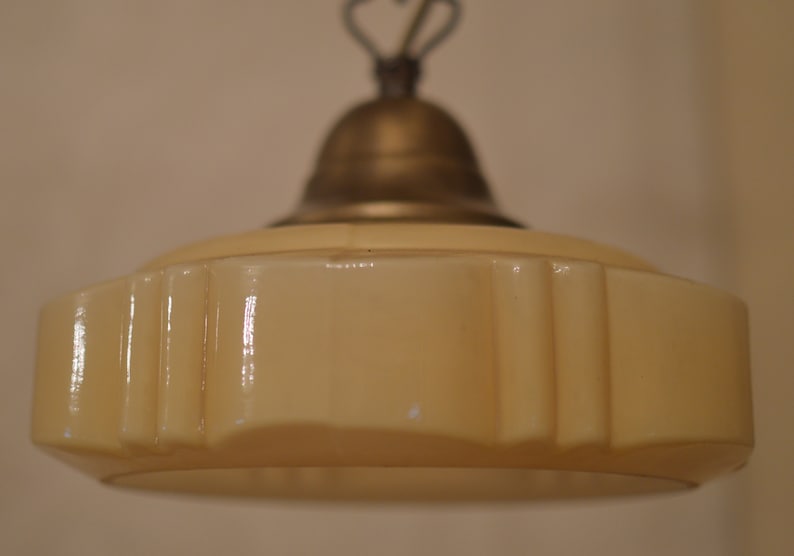 K&ouml;nnte beinhalten: Eine cremefarbene Glaspendelleuchte mit einem dekorativen, geriffelten Design. Die Leuchte hat ein Messing-Oberteil und eine Kette.