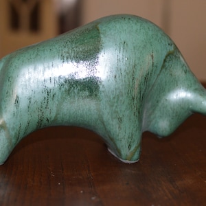 Könnte beinhalten: Eine grüne Keramik-Stierfigur mit glatter, glänzender Oberfläche. Der Stier steht auf einer Holzoberfläche.