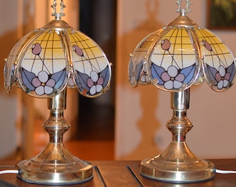 Può includere: Due lampade da tavolo dorate con paralumi in vetro colorato che presentano un motivo floreale in tonalità di giallo, blu e rosa. Le lampade sono su un tavolo di legno.