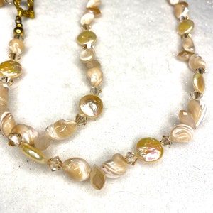 May include: A delicate necklace featuring iridescent, cream-colored shell beads and clear crystal accents. The necklace has a gold-colored clasp and is displayed on a white surface. The beads vary in shape and size, creating a unique, organic design.