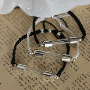 Pode incluir: Três pulseiras de amizade pretas e brancas com frascos de vidro prateados contendo mensagens escritas à mão. As pulseiras estão em um livro com texto.