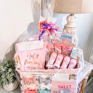 May include: A pink and white baby gift basket filled with items such as a pink onesie with the text "i got it from my mama", pink socks, a pink and gray elephant bib with the text "Sweet Little Girl", and pink and gray hair bows.