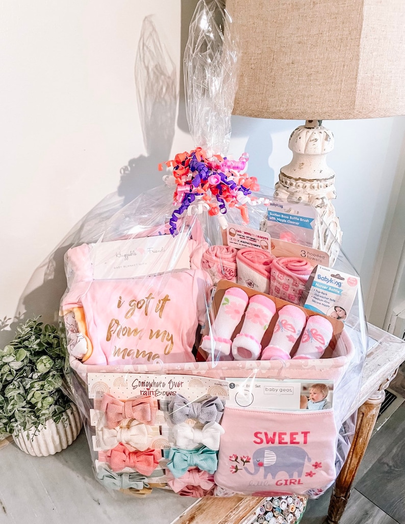 May include: A pink and white baby gift basket filled with items such as a pink onesie with the text "i got it from my mama", pink socks, a pink and gray elephant bib with the text "Sweet Little Girl", and pink and gray hair bows.