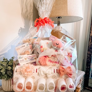 May include: A pink and white baby gift basket with a unicorn plush toy, a pink blanket, and a variety of baby items. The basket is wrapped in clear cellophane and tied with a pink ribbon.
