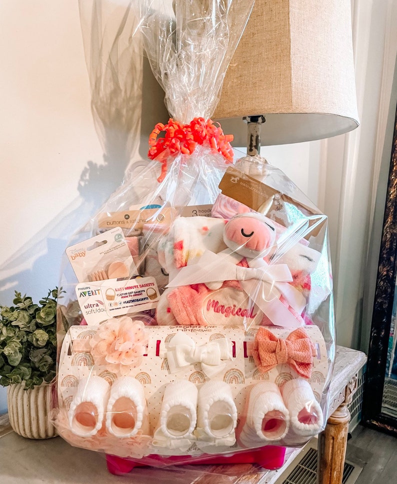 May include: A pink and white baby gift basket with a unicorn plush toy, a pink blanket, and a variety of baby items. The basket is wrapped in clear cellophane and tied with a pink ribbon.