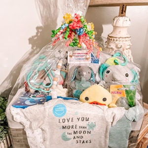 May include: A wicker basket filled with baby gifts, including a gray onesie with the text "Love you more than the moon and stars", a yellow duck toy, a green and blue rattle, and a blue and white striped blanket.