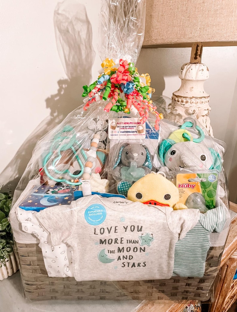 May include: A wicker basket filled with baby gifts, including a gray onesie with the text "Love you more than the moon and stars", a yellow duck toy, a green and blue rattle, and a blue and white striped blanket.