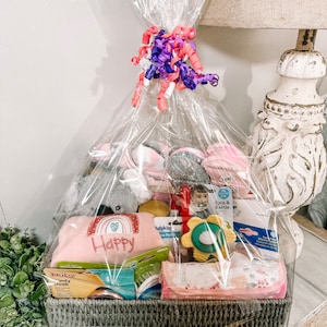 May include: A gray wicker basket filled with baby items, including a plush gray koala, a pink and white blanket with the word "Happy" embroidered on it, a yellow and green baby toy, and a pink and white baby blanket. The basket is wrapped in clear cellophane and tied with a pink and purple ribbon.