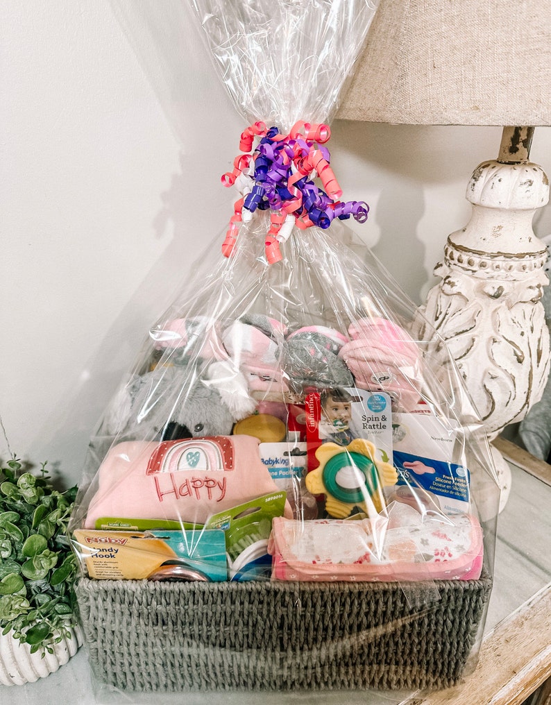 May include: A gray wicker basket filled with baby items, including a plush gray koala, a pink and white blanket with the word "Happy" embroidered on it, a yellow and green baby toy, and a pink and white baby blanket. The basket is wrapped in clear cellophane and tied with a pink and purple ribbon.