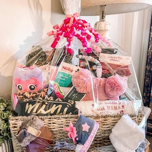 May include: A wicker basket filled with baby gifts, including a pink and purple stuffed animal, a white onesie with the text "cuddle with mama", a pair of pink and blue baby shoes, and a pink and white blanket. The basket is wrapped in clear cellophane and tied with a pink ribbon.