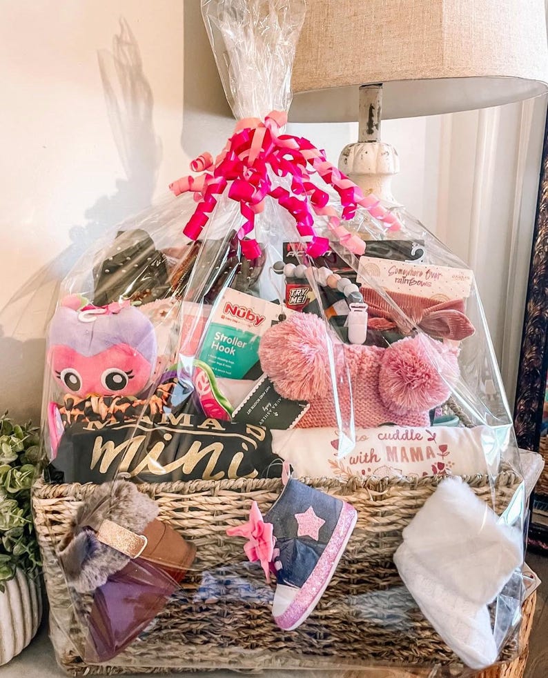 May include: A wicker basket filled with baby gifts, including a pink and purple stuffed animal, a white onesie with the text "cuddle with mama", a pair of pink and blue baby shoes, and a pink and white blanket. The basket is wrapped in clear cellophane and tied with a pink ribbon.