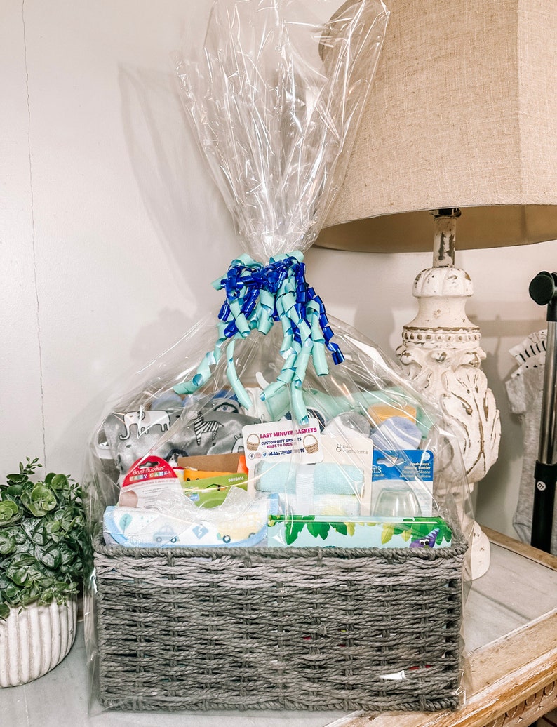 May include: A gray wicker basket filled with baby items, including a blue and white patterned blanket, a stuffed animal, and a bottle of baby lotion. The basket is wrapped in clear cellophane and tied with a blue and green ribbon. The text "Last Minute Jackets" is visible on a tag attached to the basket.